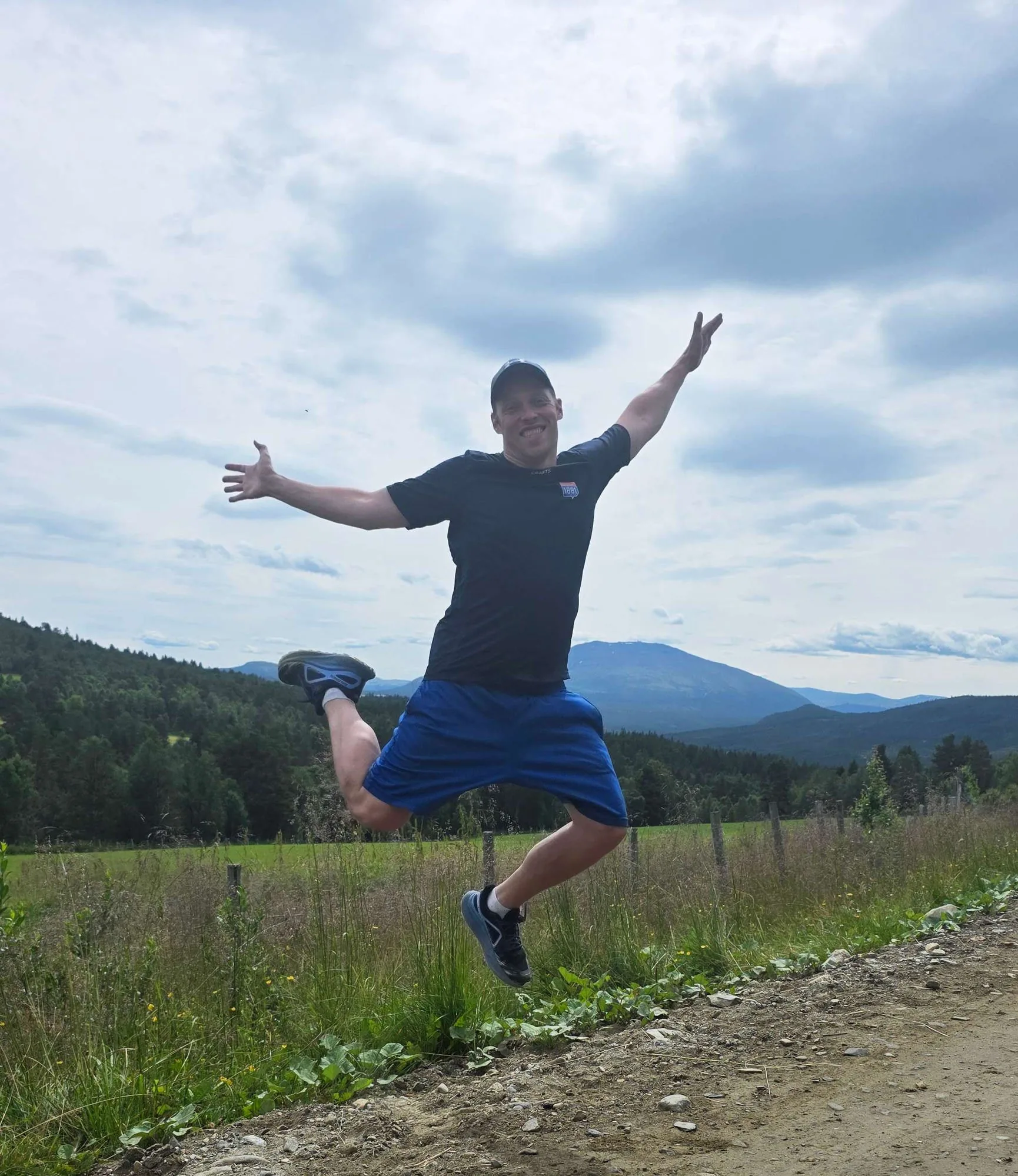 Magnus mid-jump on a gravel road, arms wide, with Norwegian forest and mountains behind him